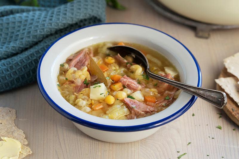 Ertesuppe med grønnsaker og saltet svineknoke, servert i hvit suppeskål med mørk blå kant, på lyst trebord. Mørkt blått kjøkkenhåndkle, og flattbrød ved siden av.