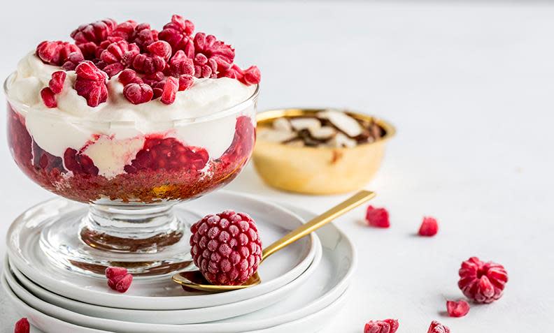 Bringebærsurprise; dessert med krem, vaniljeis, knuste kjeks og bringebær, servert i dessertglass på hvit skål. Liten gullskål ved siden av med skivede mandler, og ferske bringebær strødd rundt.