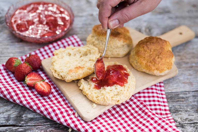 Hjemmelagede scones servert på trefjøl med rødrutete serviett under. En av sconesene er delt i to, og en hånd legger jordbærsyltetøy på en av halvdelene.