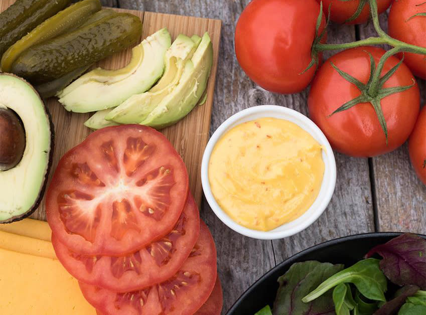 Liten hvit skål med hjemmelaget chilimajones, på rustikk trebord, omringet av ferske tomater, avokado, fersk salat, pickles og burgerost.