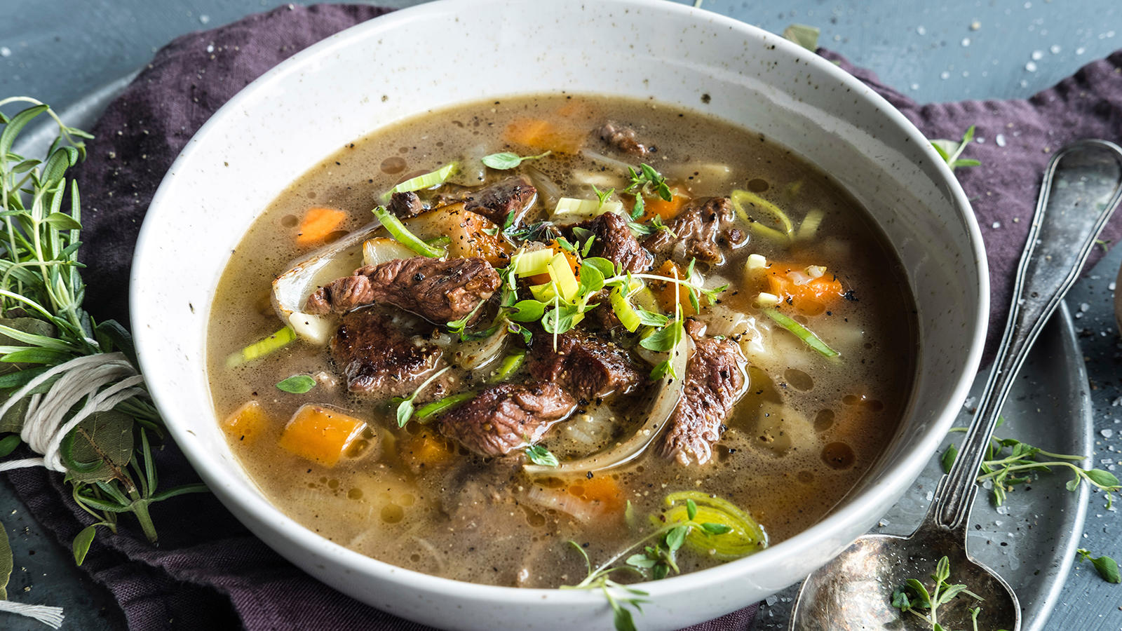 Kjøttsuppe med høyrygg og rotgrønnsaker servert i lys keramikkbolle med friske urter på toppen. Et knytte med friske urter og en gammeldags suppeskje på siden.