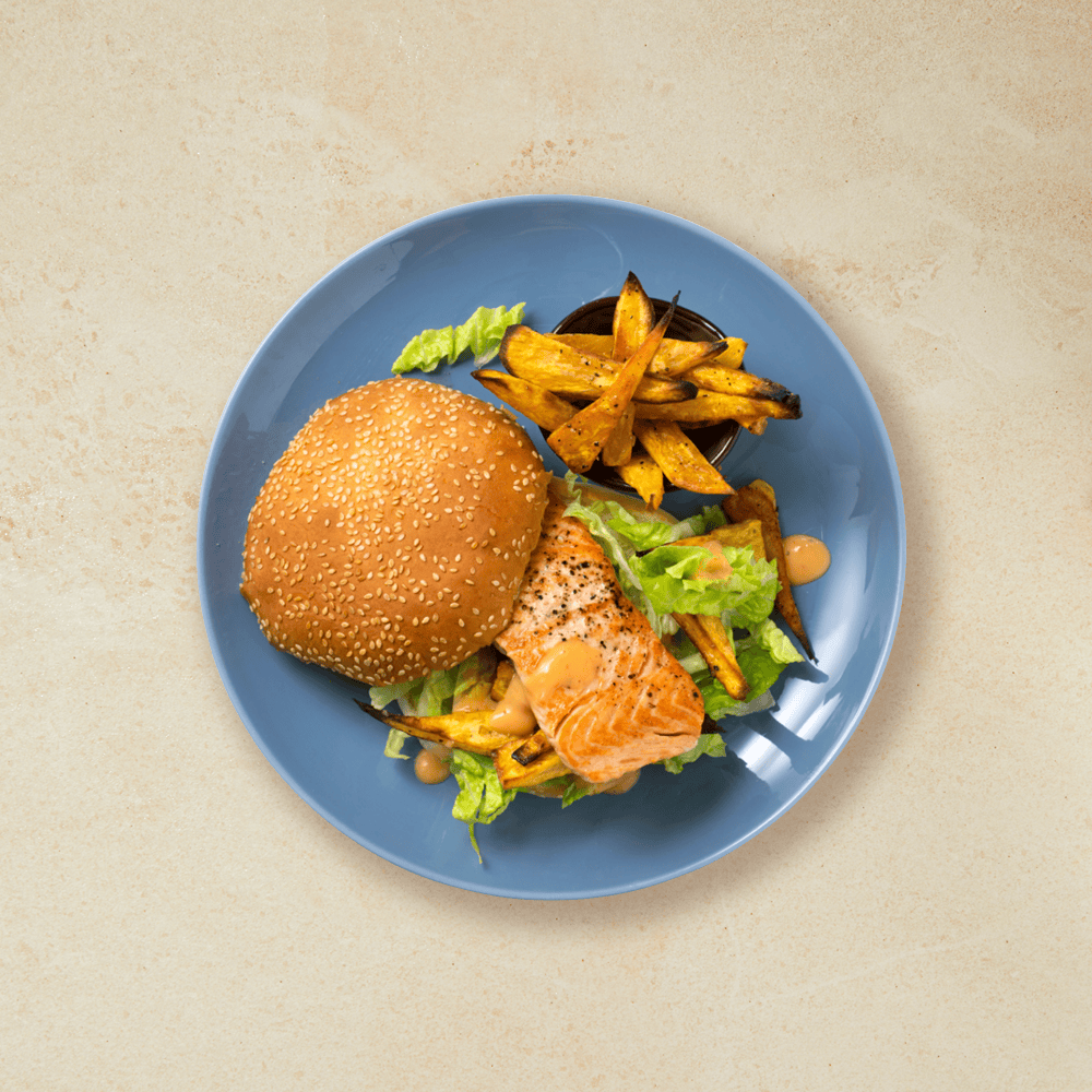 Stekt lakseburger av laksefilet, servert i burgerbrød med salat, dressing og hjemmelagede søtpotetfries på blå tallerken.