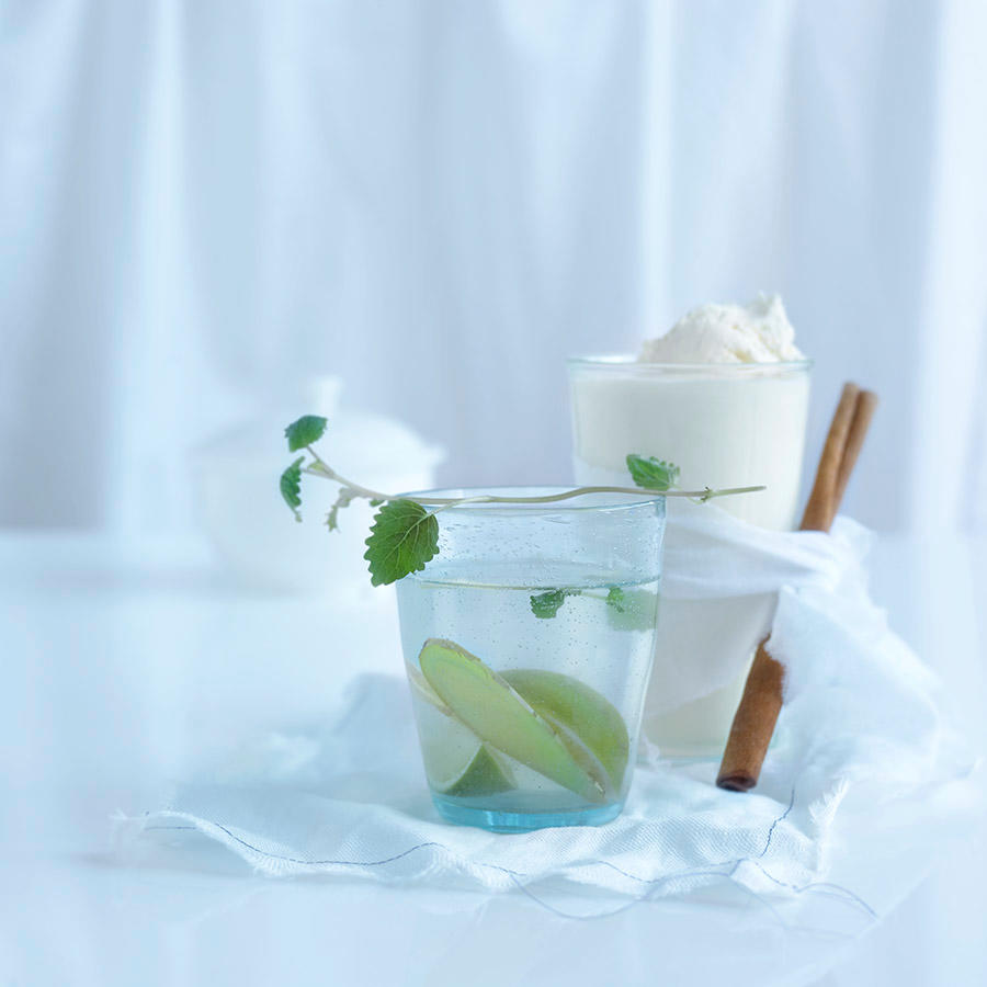 Ingefærte med honning og lime servert i kjøkkenglass på hvit tøyserviett på hvitt bord. En kvist fersk mynte på toppen.