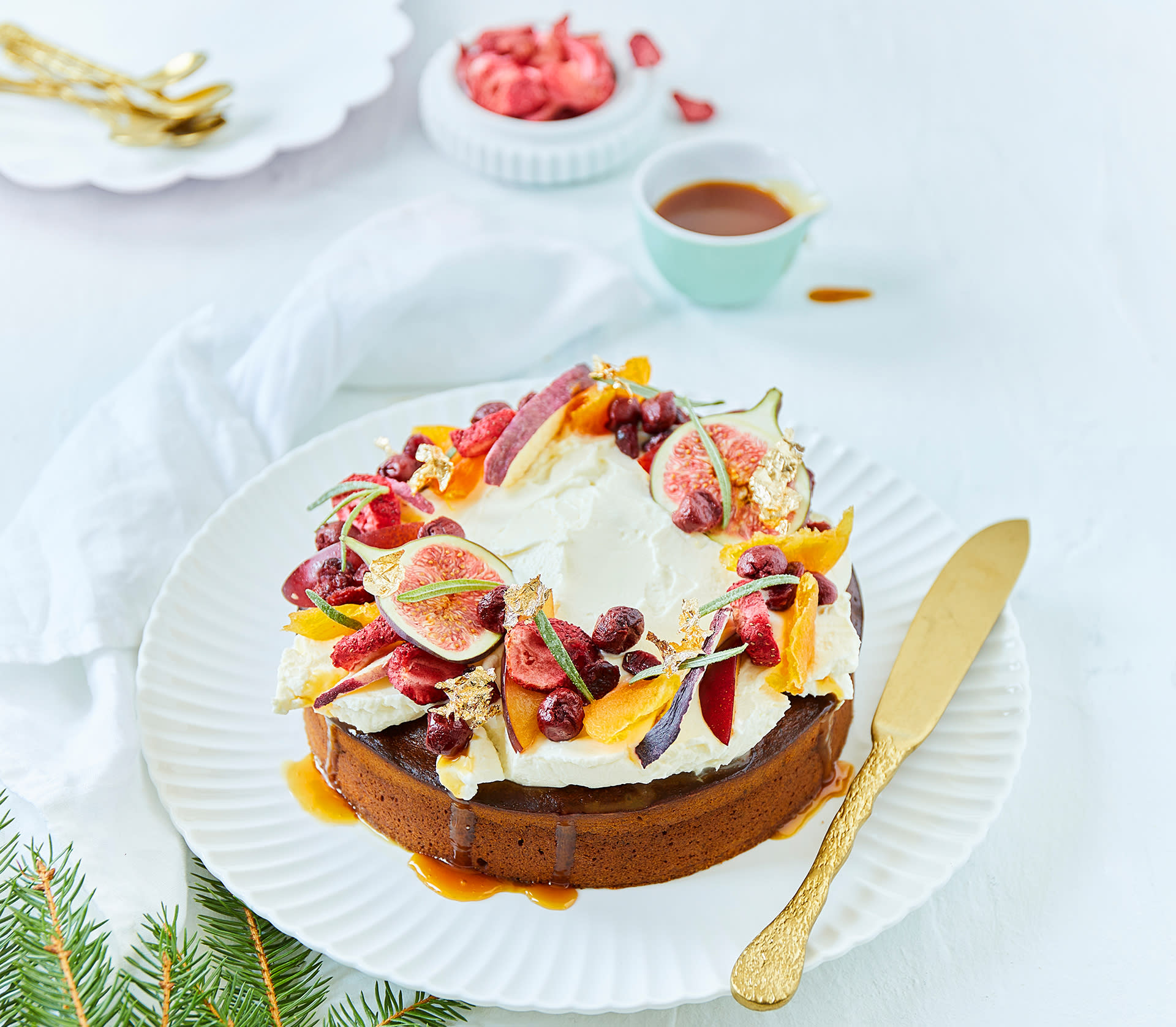 Daddelkake med mascarpone og salt karamell, pyntet med tørkede frukt og bær, ferske epler i skiver, og halve fiken, servert på hvitt rundt fat med gullkniv på kanten.