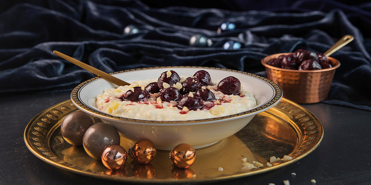 Riskrem med marsipan og kirsebær servert i gullkantet hvit dessertskål på gullfarget dekketallerken, med små julekuler rundt. Liten messingskål med kirsebær i bakgrunnen.