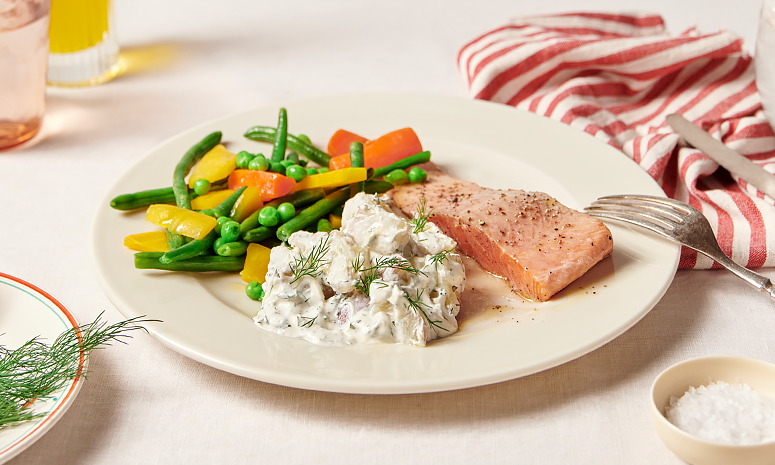 Ørret og poteter med rømme og dill servert på en hvit tallerken med en gaffel liggende på høre siden, et rødt og hvitt stripete håndkle ligger på høre siden i bakgrunnen