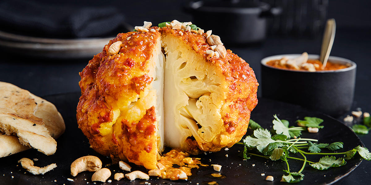 Gratinert blomkålhode med spicy indisk saus servert på svart tallerken med nanbrød, cashewnøtter og frisk koriander. Skål med indisk saus i bakgrunnen.