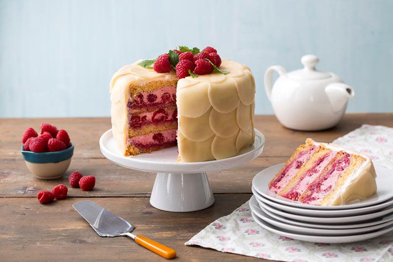 Marsipankake med bringebærkrem inni, pyntet med sirkler av marsipan, ferske bringebær og sitronmelisse, servert på hvitt stettfat på rustikk trebord.