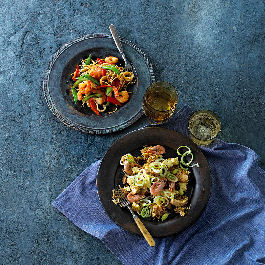 Scampi teriyaki med nudler og grønnsaker servert på mørk tallerken, på mørk bakgrunn ved siden av tallerken med Nasi Goreng.