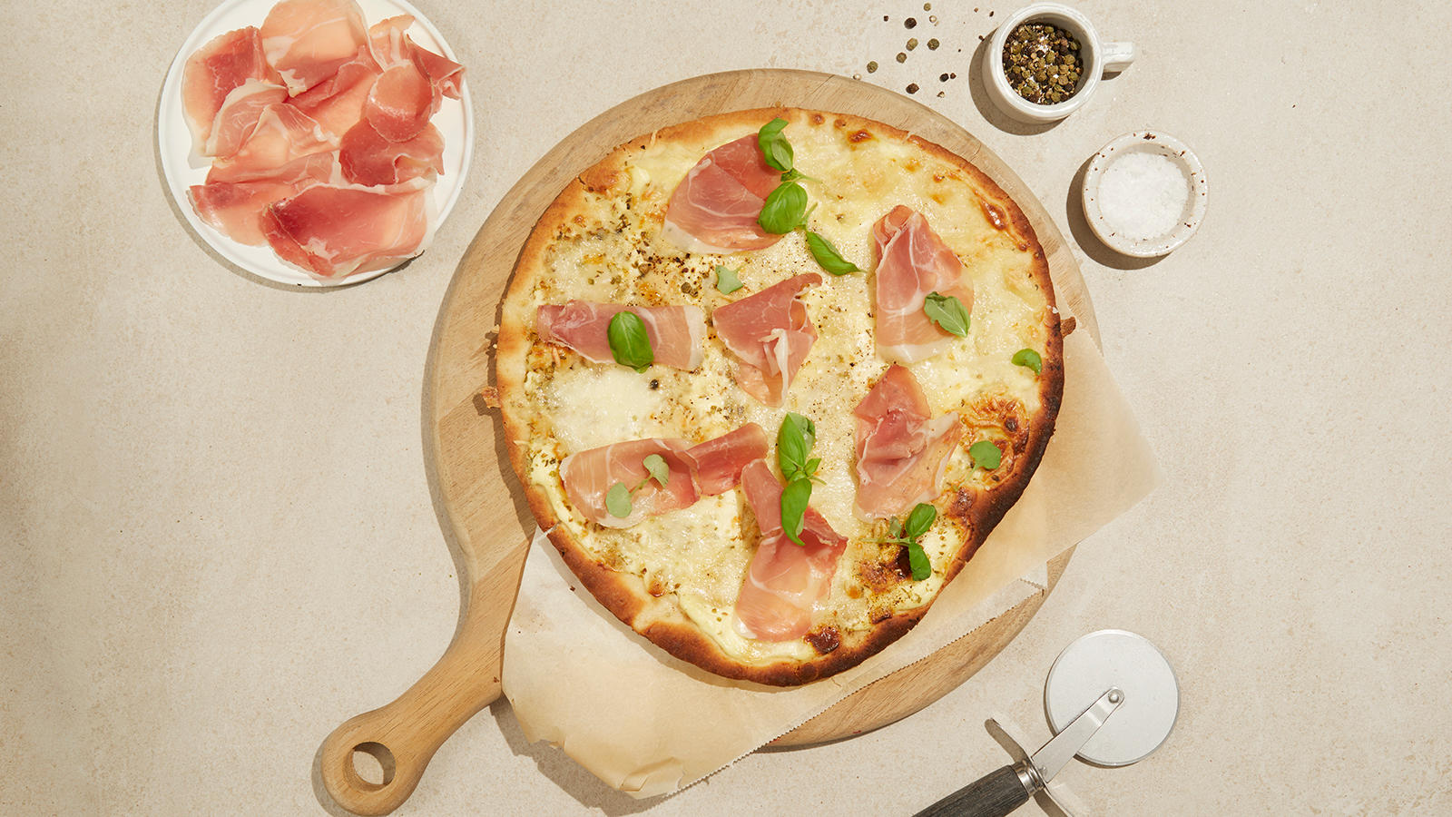 Hvit pizza med spekeskinke servert med frisk basilikum på lys trefjøl. Lite fat med spekeskinke ved siden av.