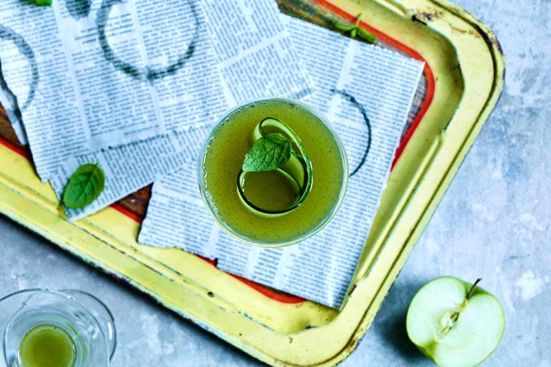 Grønn drink med agurk- og myntepynt på serveringsbrett med avispapir og oppkuttede epler rundt