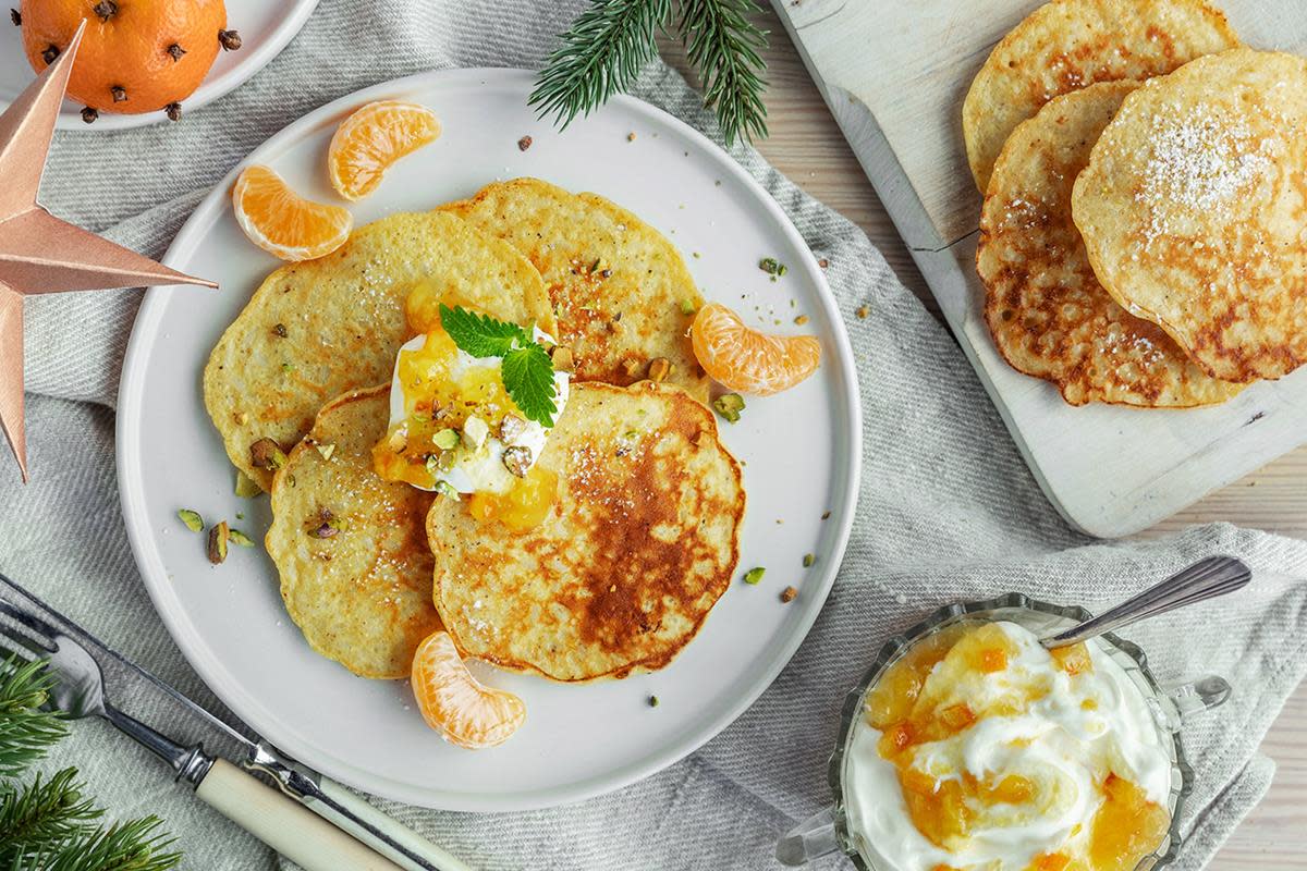 Gyldne rislapper servert på hvit tallerken, med rømme, klementinbåter og et dryss pistasjenøtter. Står på julepyntet bord i lyse og beige farger. Klementin med nellikspiker ved siden av.