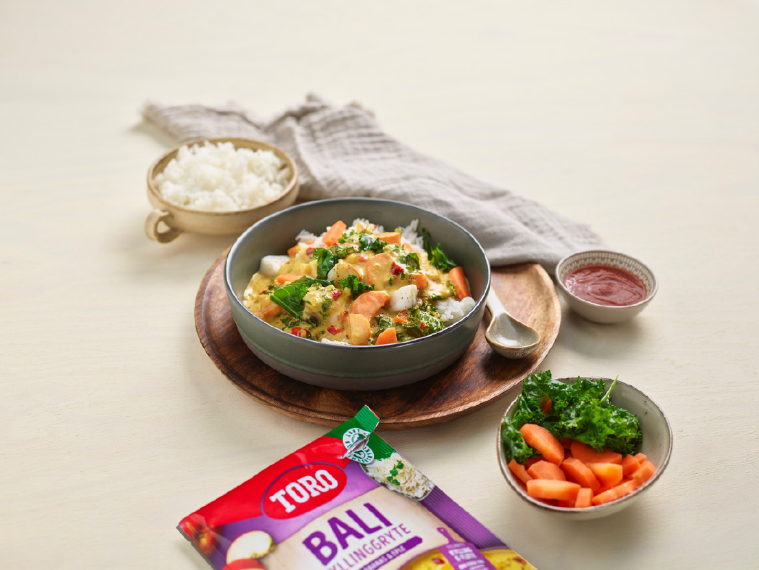 Baligryte med torsk i en skål på en tretallerken. Til venstre i bakgrunnen er det en skål med ris og til høyre er det en skål mer sriracha og en skål med gulrøtter og grønnkål. I forgrunnen ligger en Toro Baligryte pose