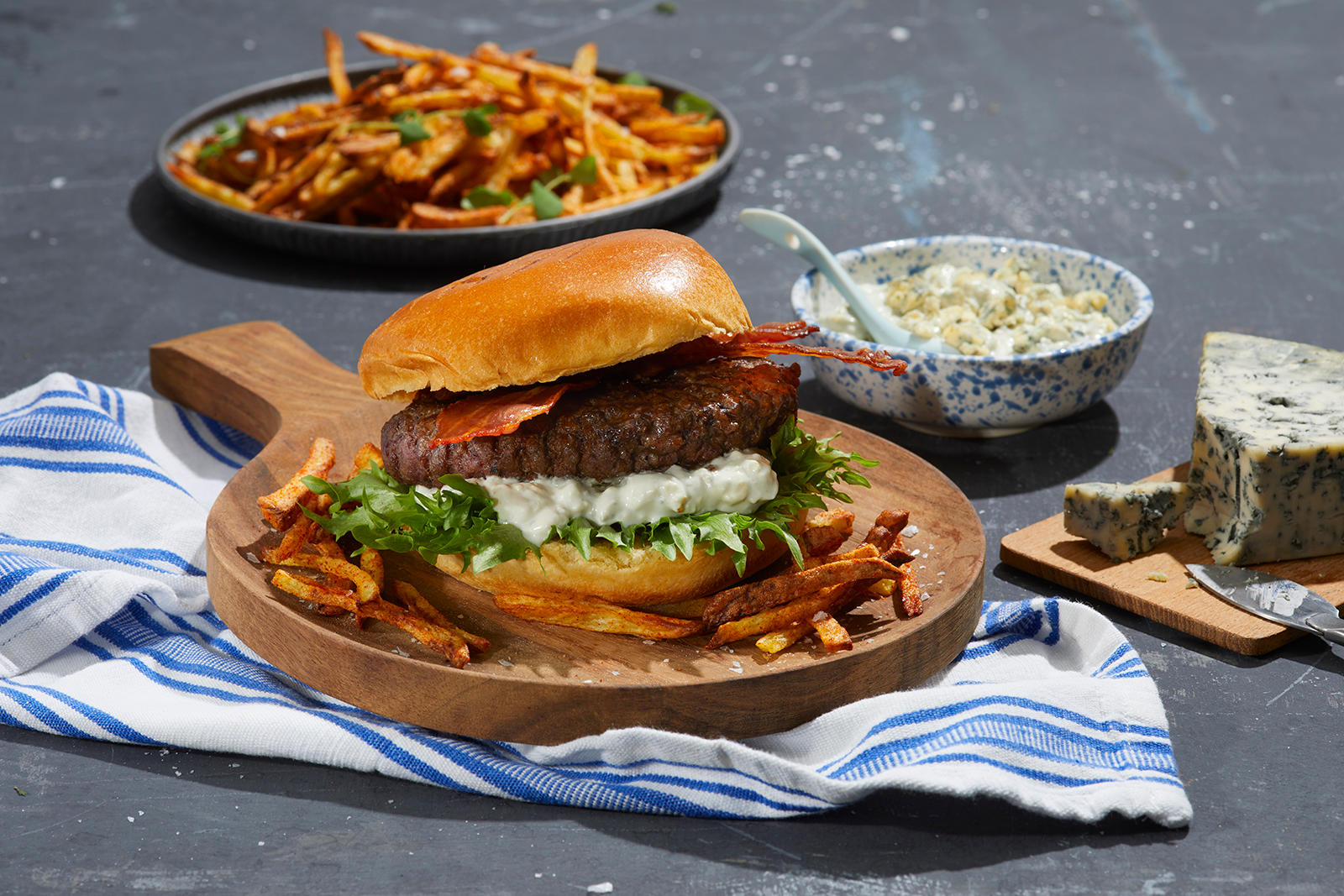 Big Juicy burger med blåmuggost og skinnyfries, servert med bacon og salat på rund trefjøl på grått steinunderlag. Liten fjøl med blåmuggost, og blåmønstret skål med blåmuggostdressing ved siden av. Fat med skinnyfries i bakgrunnen.