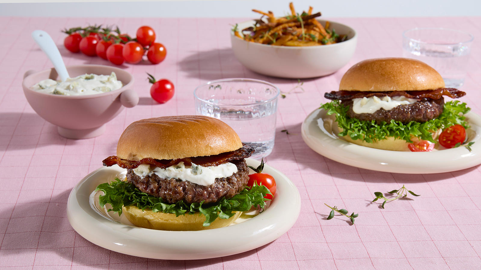 Big Juicy burger med blåmuggostdressing og skinnyfries, servert med bacon og salat på lyse tallerkner på rosa rutet duk.