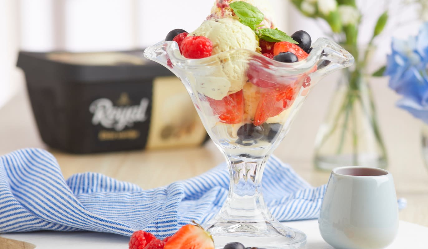 Iskrem med hjemmelaget jordbærsaus servert i iskremglass med friske bringebær, blåbær og jordbær. Glasset står på en hvit trefjøl på et lyst bord med blå blomster i bakgrunnen og en boks Royal vaniljeis på siden.