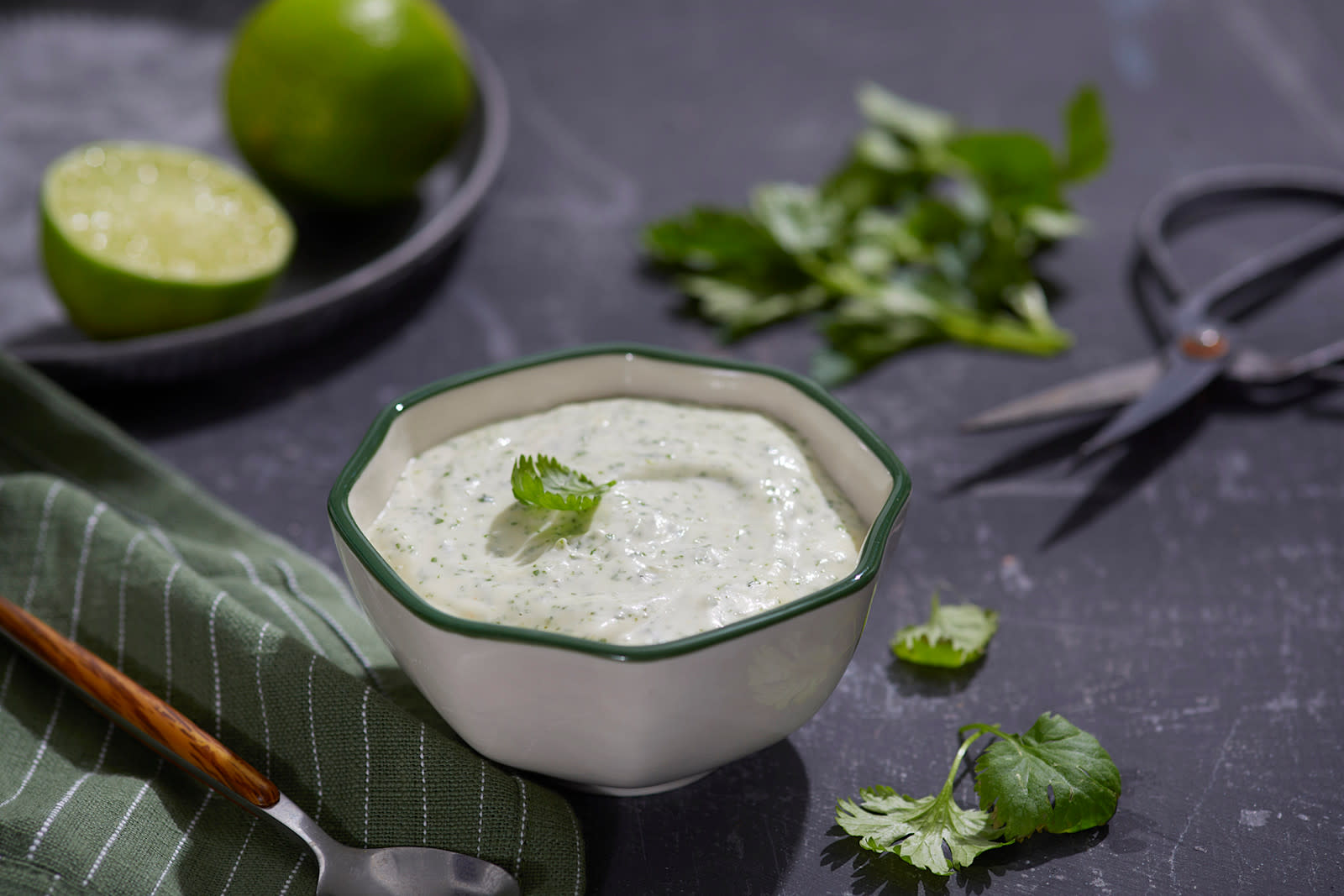 Hjemmelaget aioli med koriander servert i liten hvit skål med grønn kant, på mørkegrå steinbakgrunn. Frisk koriander og urtesaks ved siden av, og hel og halv lime i bakgrunnen.