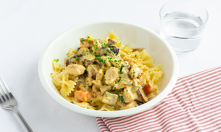 Pasta med kylling, rotgrønnsaker og sopp i fløtesaus pyntet med gressløk i en hvit skål på et rødt og hvitt stripete håndkle.