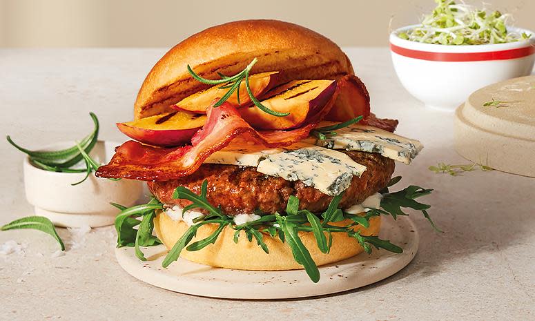 Burger med bacon, grillet fersken og blåmuggost, på liten beige tallerken. Lys bakgrunn.