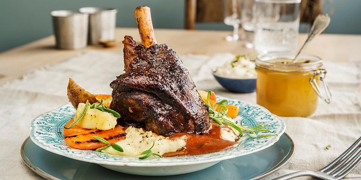 Braisert lammeskank med hjemmelaget potetmos, smørstekte rotgrønnsaker og pepperrotkrem servert på blåmønstret asjett, med dekket bord i bakgrunnen.