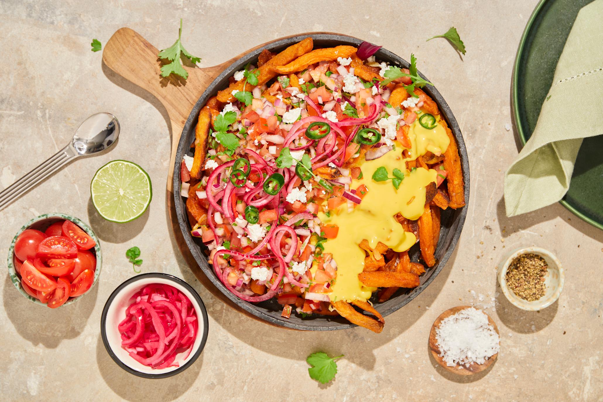 Dirty søtpotetfries med pico de gallo og Kavli cheddar dipp servert i rund ildfast form på trefjøl, og toppet med syltet rødløk, grønn chili og frisk koriander. Små skåler med syltet rødløk og tomat på siden.