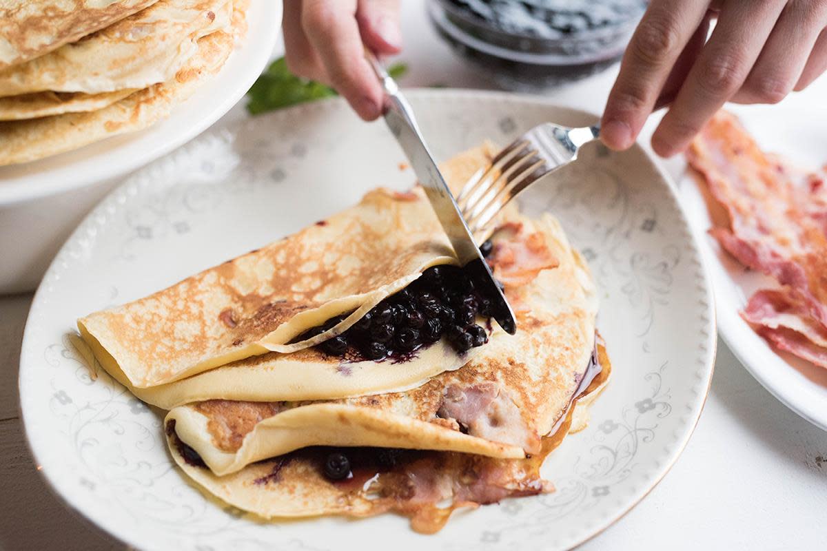 Fleskepannekaker på middagstallerken, toppet med blåbærsyltetøy. To hender holder bestikk som skjærer en bit av den øverste pannekaken. Stekt bacon ligger ved siden av tallerkenen.