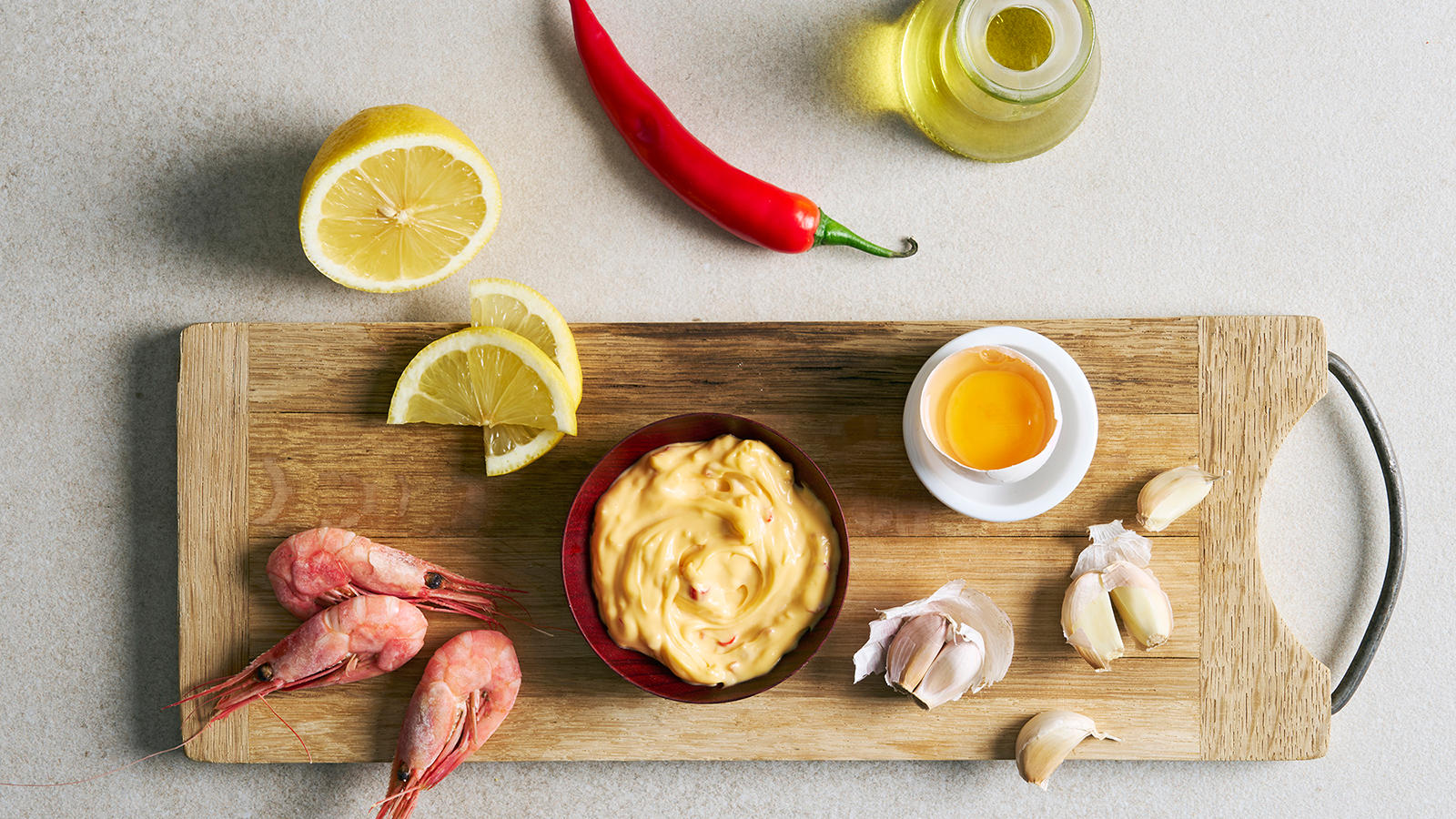 Chilimajones med hvitløk servert i liten skål på trefjøl med hel chili, sitronskiver, eggeplomme, hvitløksfedd og hele reker ved siden av.