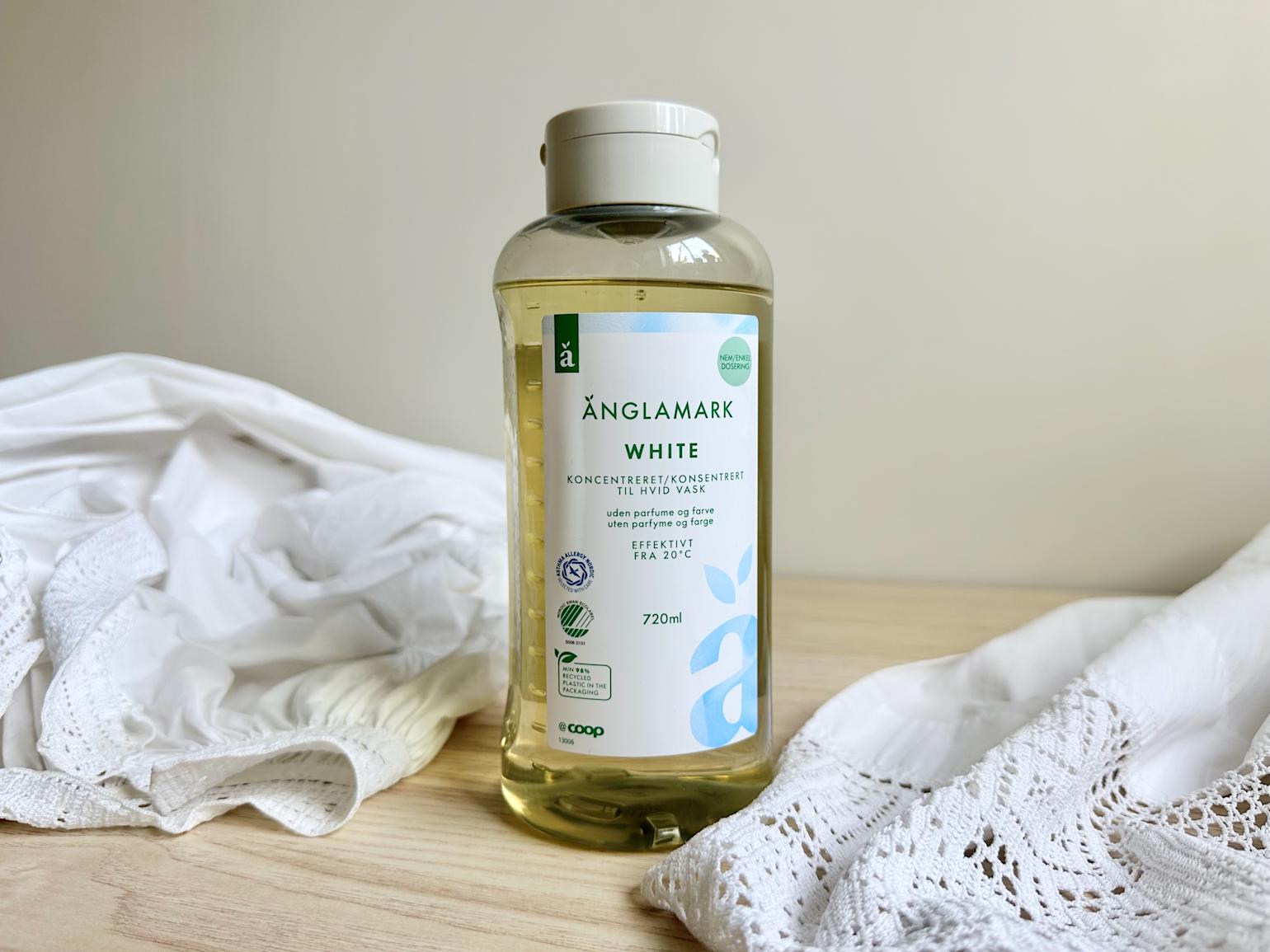 Bildet viser en flaske Änglamark White vaskemiddel stående på en lys treflate, omgitt av en hvit bunadsskjorte med broderidetaljer. Produktet er svanemerket og utviklet for skånsom og effektiv vask av hvitt tøy – perfekt for delikate tekstiler som bunadsskjorter.