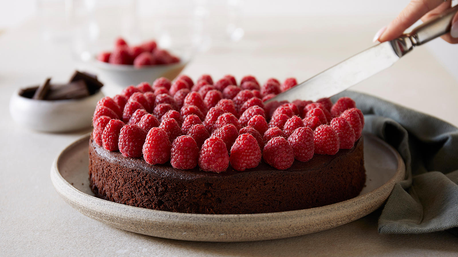 Glutenfri sjokoladekake uten melk pyntet med friske bringebær og servert på keramikkasjett. To små skåler i bakgrunnen med høvlet sjokolade og friske bringebær. Hånd skjærer en bit av kaken med sølvkniv.