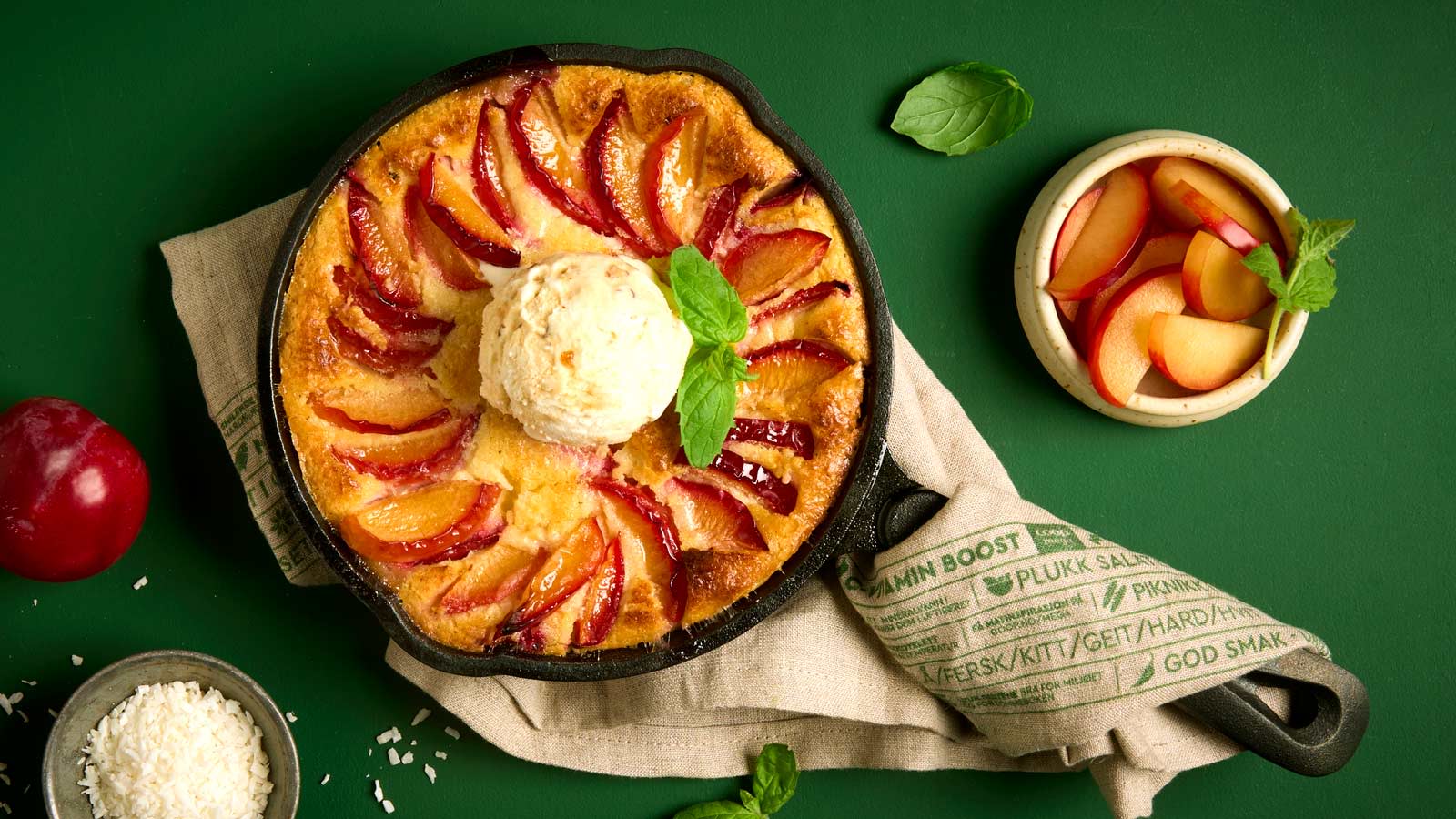 Plommeclafoutis servert i svart støpejernspanne med en kule is og grønne urter på toppen. Liten skål friske røde plommer på siden.
