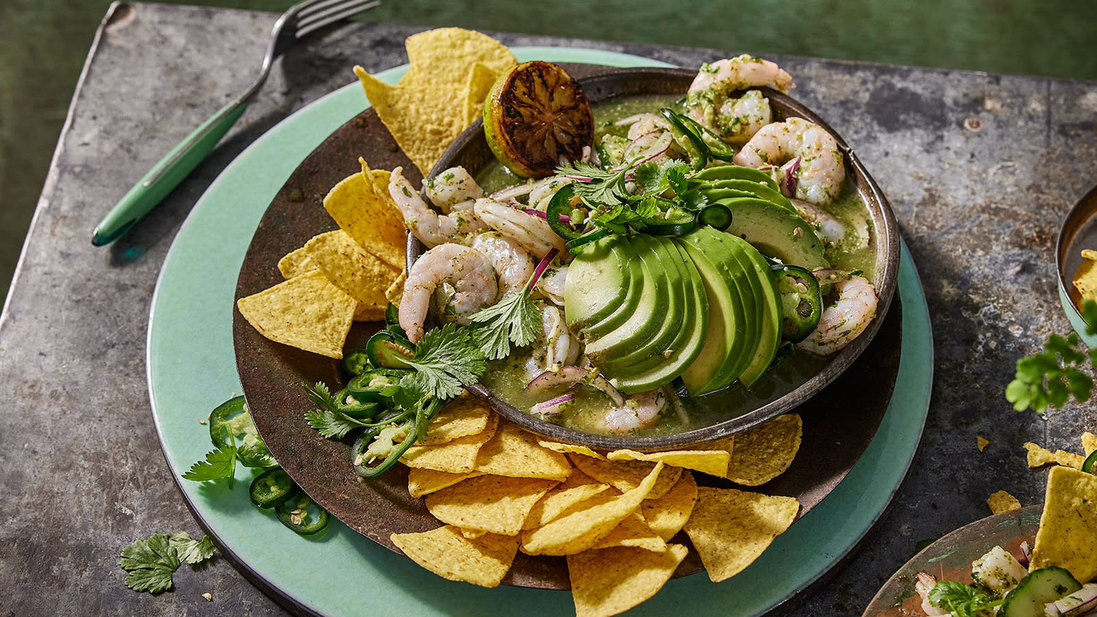 Lime- og chilimarinerte scampi servert med avokadoskiver, tortillachips og koriander i mørk skål på turkis asjett.