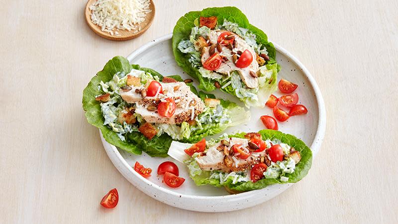 Kyllingsalat med parmesan, tomater og ristede gresskarkjerner, servert i hjertesalatblader på hvit tallerken. Lys bakgrunn.