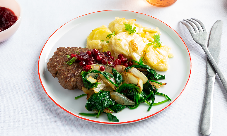 Karbonader med fløtegratinerte poteter og stekt løk og grønnsaker toppet med tyttebærsyltetøy servert på en hvit tallerken med kniv og gaffel på høyre sided