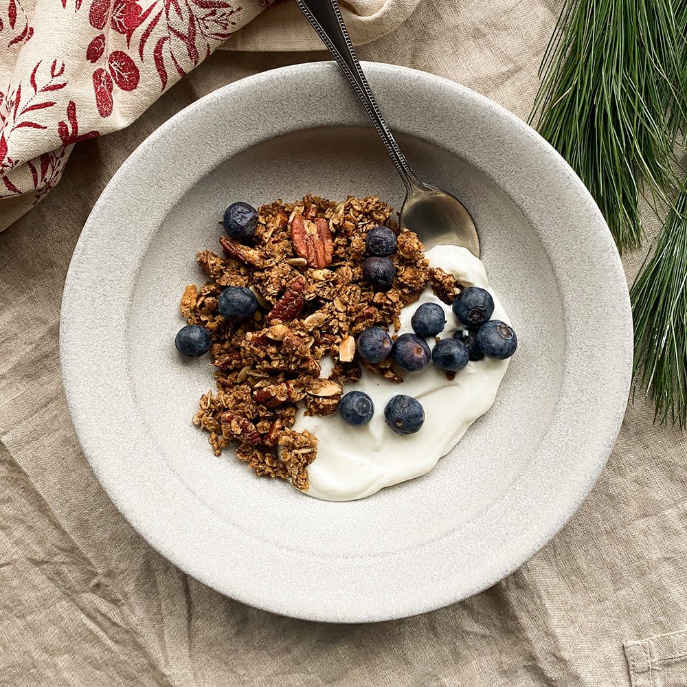 Pepperkakegranola med nøtter servert over gresk yoghurt i lys skål med friske blåbær. Granbar og juleduk i lin ved siden av.