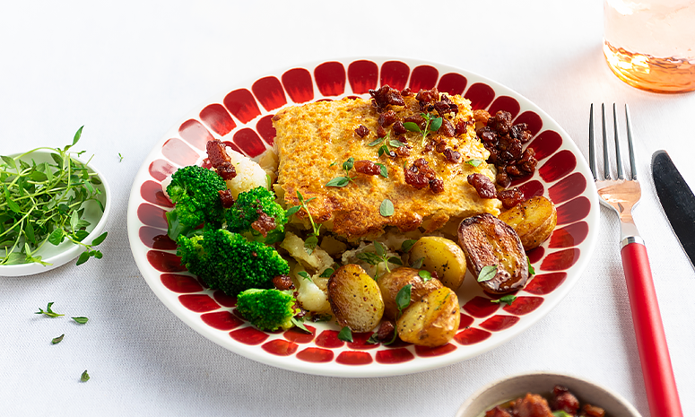 Fiskegrateng med poteter og brokkoli på rød tallerken pyntet med urter fra en hvit utreskål på venstre side av retten.