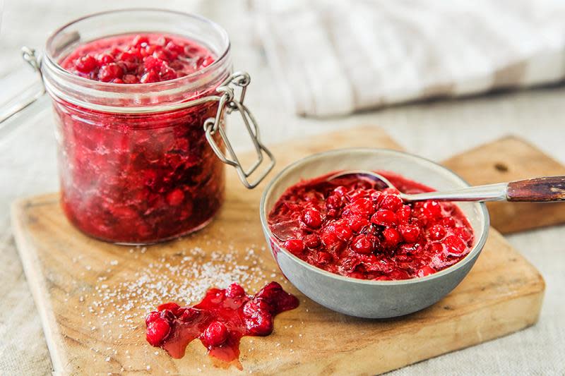 Norgesglass og keramikkskål med friskt rødt tyttebærsyltetøy, står på en skjærefjøl av tre, med en stripete linduk i bakgrunnen.