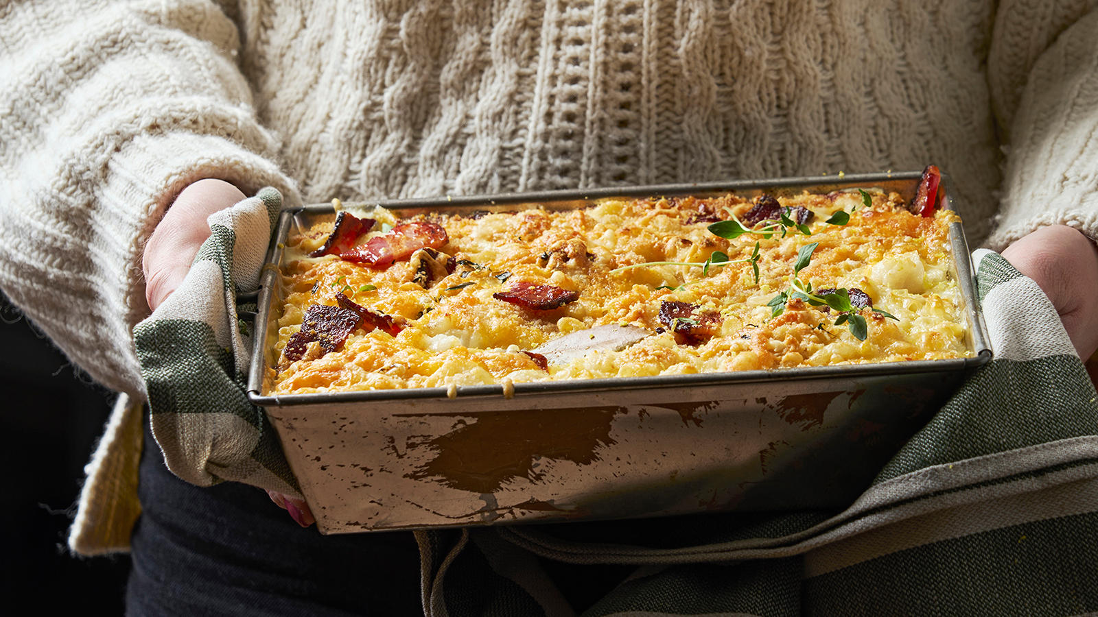 Mac and cheese med torsk og bacon servert i ildfast form av person med hvit strikkegenser.