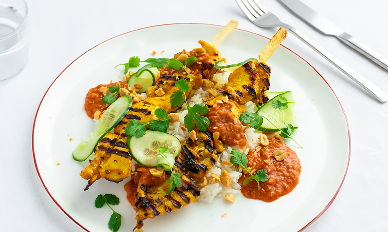 Kyllingspyd med sataysaus, peanøtter og ris toppet med tynne agurkskiver, servert på en hvit tallerken på en hvit duk med kniv og gaffel ved siden a