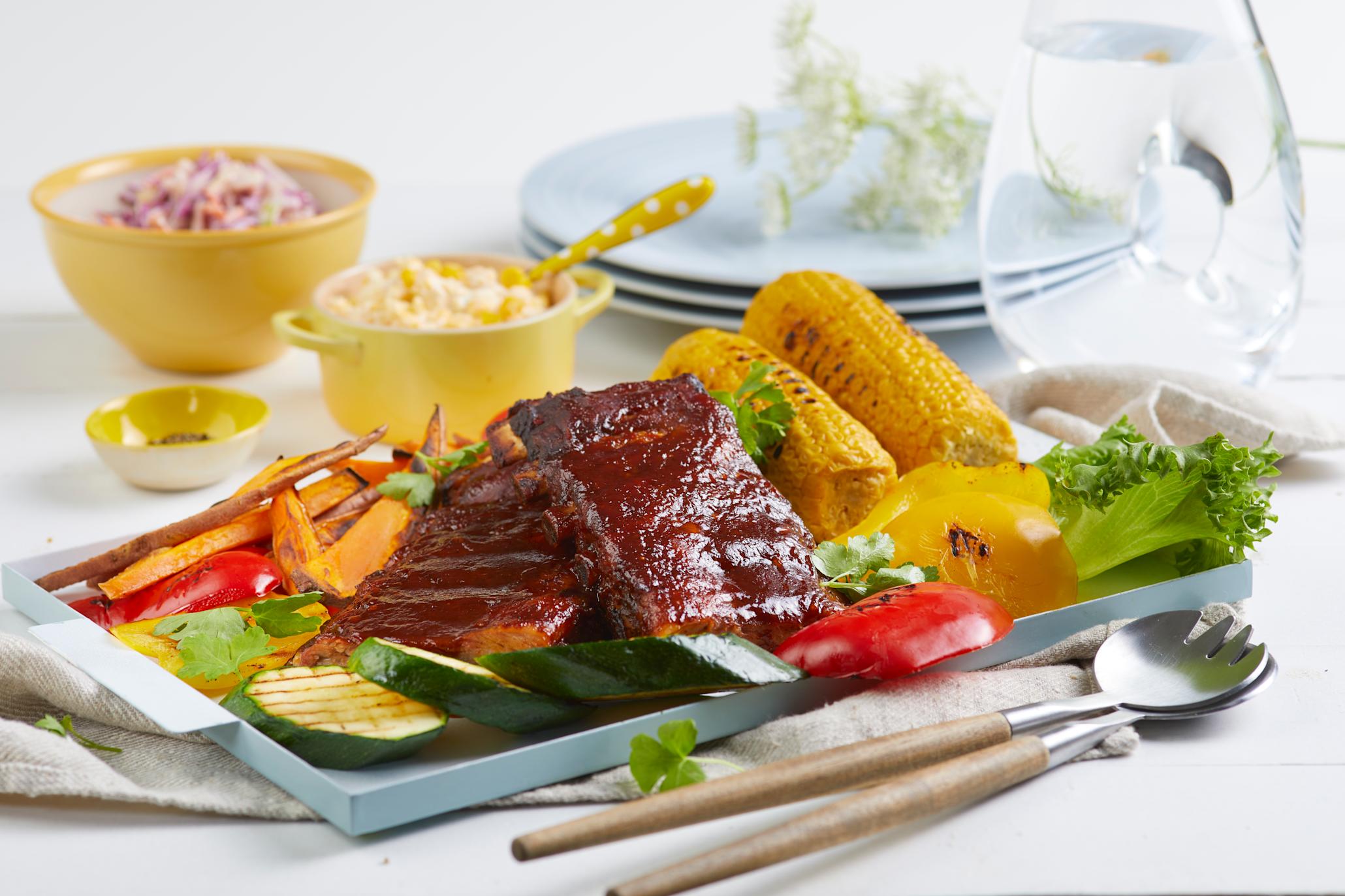 Spareribs med hjemmelaget maiskrem, ovnsbakt søtpotet og coleslaw servert på lyseblått fat med grillede grønnsaker og maiskolber. Gule skåler med coleslaw og maiskrem i bakgrunnen.