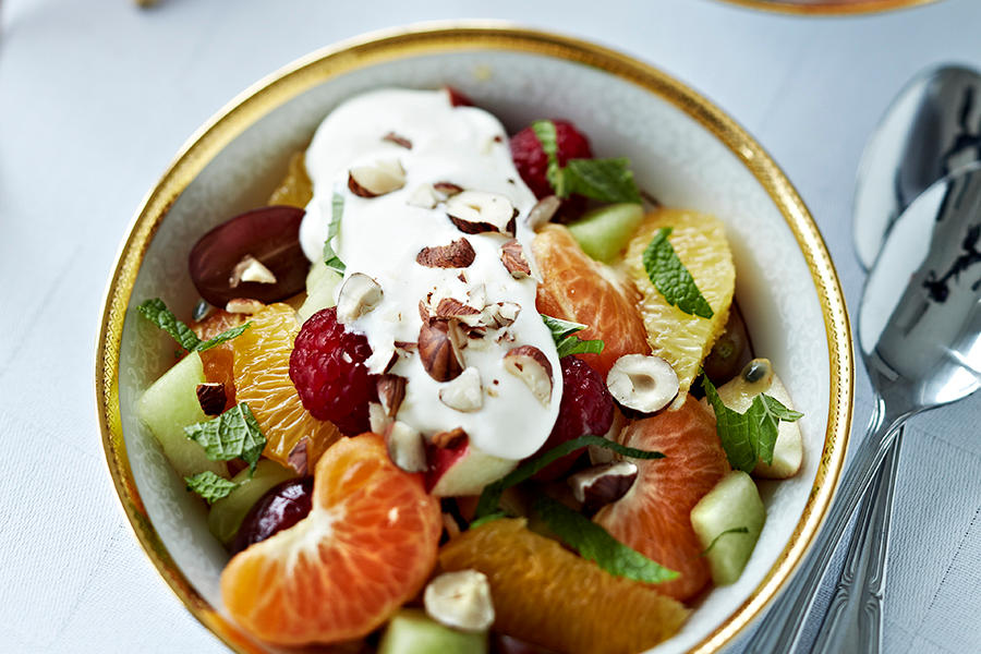 Skål med frisk fruktsalat med bringebær, klementinbåter, appelsinbåter og melon toppet med vaniljeyoghurt og hakkede hasselnøtter.