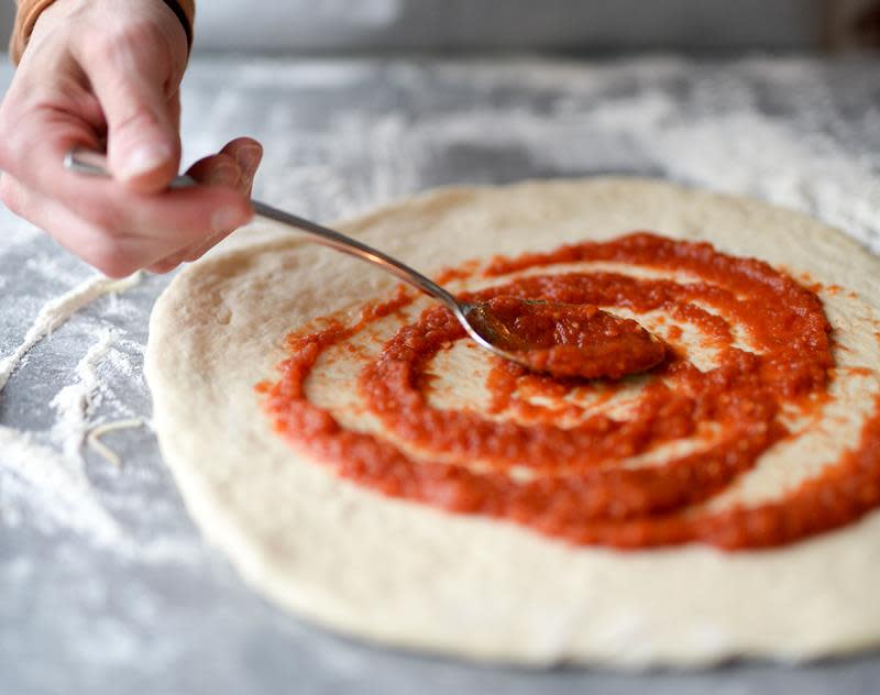 Ferdig kjevlet rund pizzadeig på melet overflate, med hånd som stryker rød pizzasaus over med en skje.