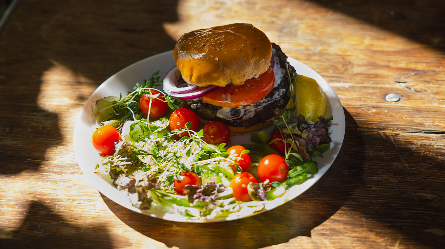 Hamburger med tomat, løk og salat på hvit tallerken, servert med frisk salat og cherrytomater på trebord.