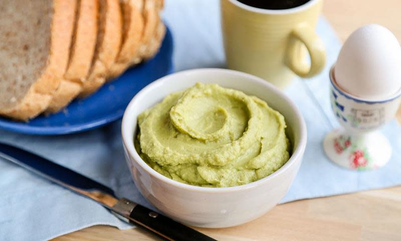 Hvit skål med avokadohummus står på frokostbord ved siden av brødskiver, en kaffekopp og et kokt egg.