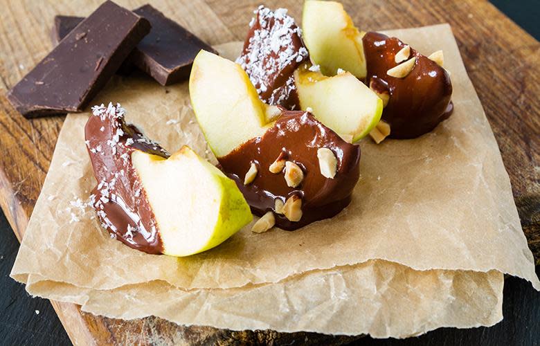 Fire båter grønt eple dyppet i mørk sjokoladefondue, og toppet med annenhver kokosmasse og hakkede nøtter. Båtene ligger på bakepapir på en trefjøl, med biter av mørk sjokolade ved siden av.