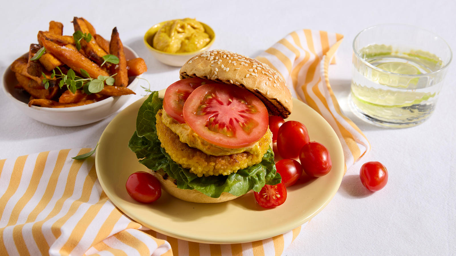 Crispy fiskeburger med currymayo og søtpotetfries servert på gul asjett med cherrytomater. Skål med søtpotetfries i bakgrunnen.