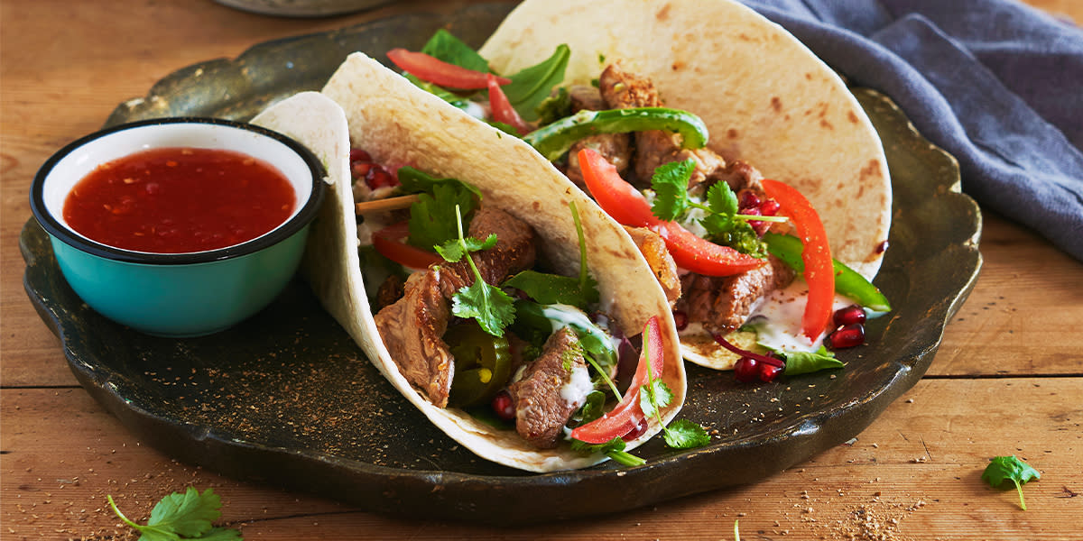 Lammetaco med salsa verde. To tacolefser fylt med stekt lammekjøtt i biter, rød og grønn paprika, koriander, granateplekjerner og en skål med rød chilisaus. 