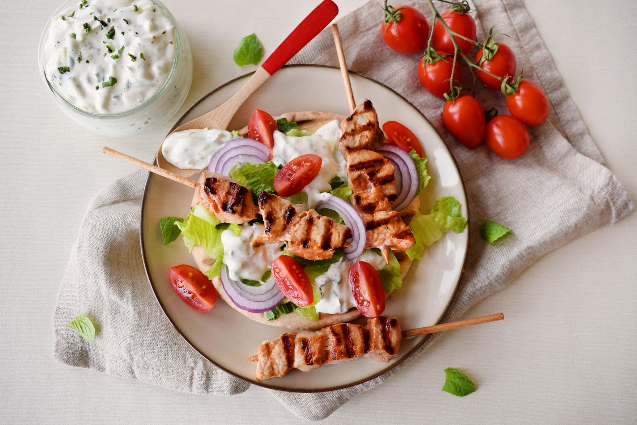 BBQ grillspyd av svin med jukse-tzatziki, servert over grillet pitabrød med tomat, rødløk og hjertesalat. Skål med jukse-tzatziki i bakgrunnen.