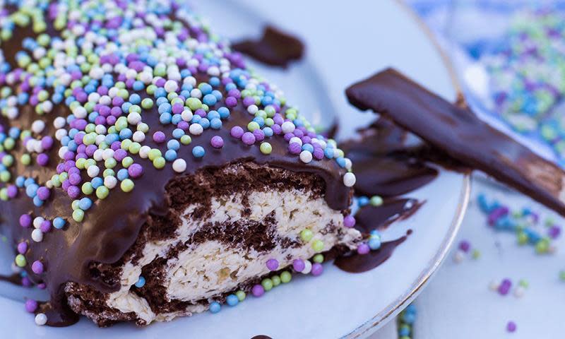Sjokoladerullekake med banankrem, sjokoladeglasur og kakestrø på toppen i lilla, hvitt, blått og grønt. Kaken står på hvitt serveringsfat.