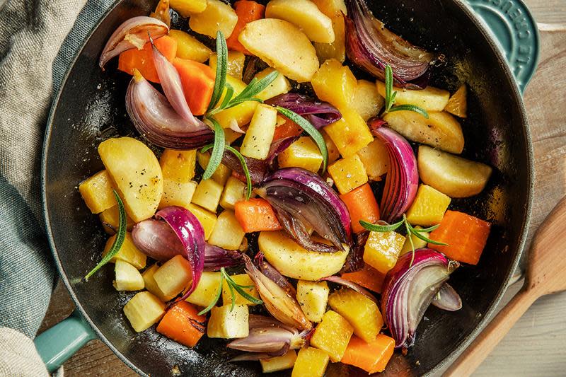 Fargerike rotgrønnsaker, eplebåter og båter av rødløk stekt i sjøgrønn jernpanne, og pyntet med fersk rosmarin. Pannen står på et trebord, med grønt og beige stripet linhåndkle.