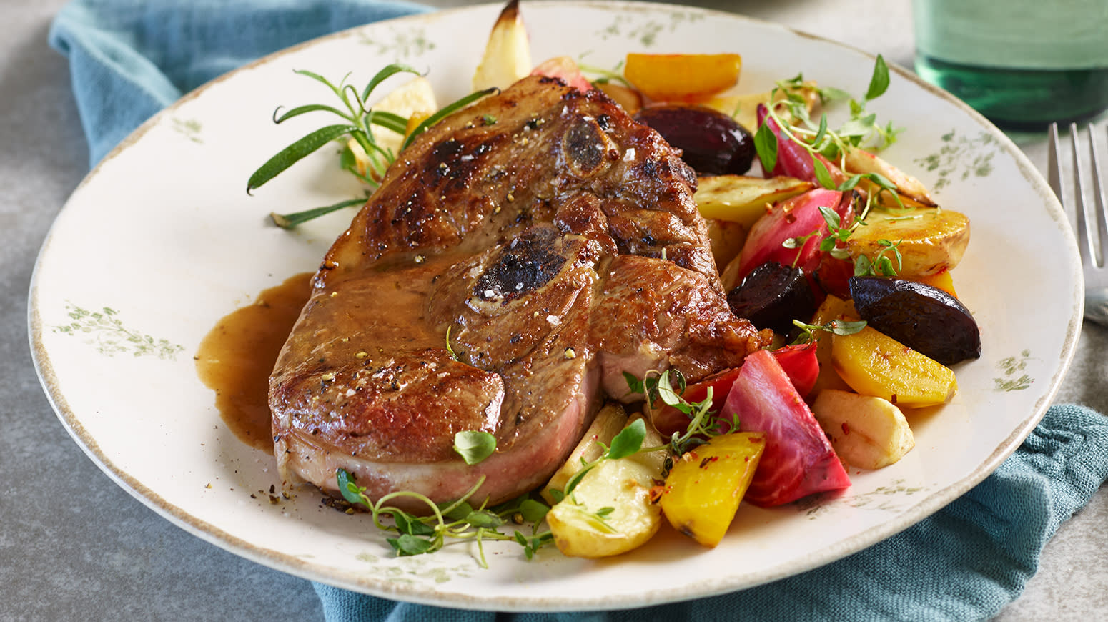 Lammelår med ovnsbakte grønnsaker, servert med friske urter på hvit tallerken med lysegrønt mønster på dekket middagsbord.